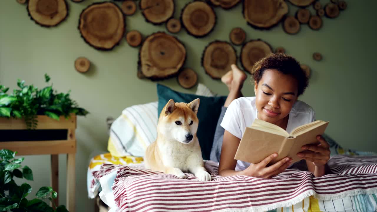 una alegre adolescente de raza mixta está leyendo un libro disfrutando de la literatura y luego acariciando a su perro shiba inu acostado en la cama cerca de ella. pasatiempo, estilo de vida moderno y concepto de animales.