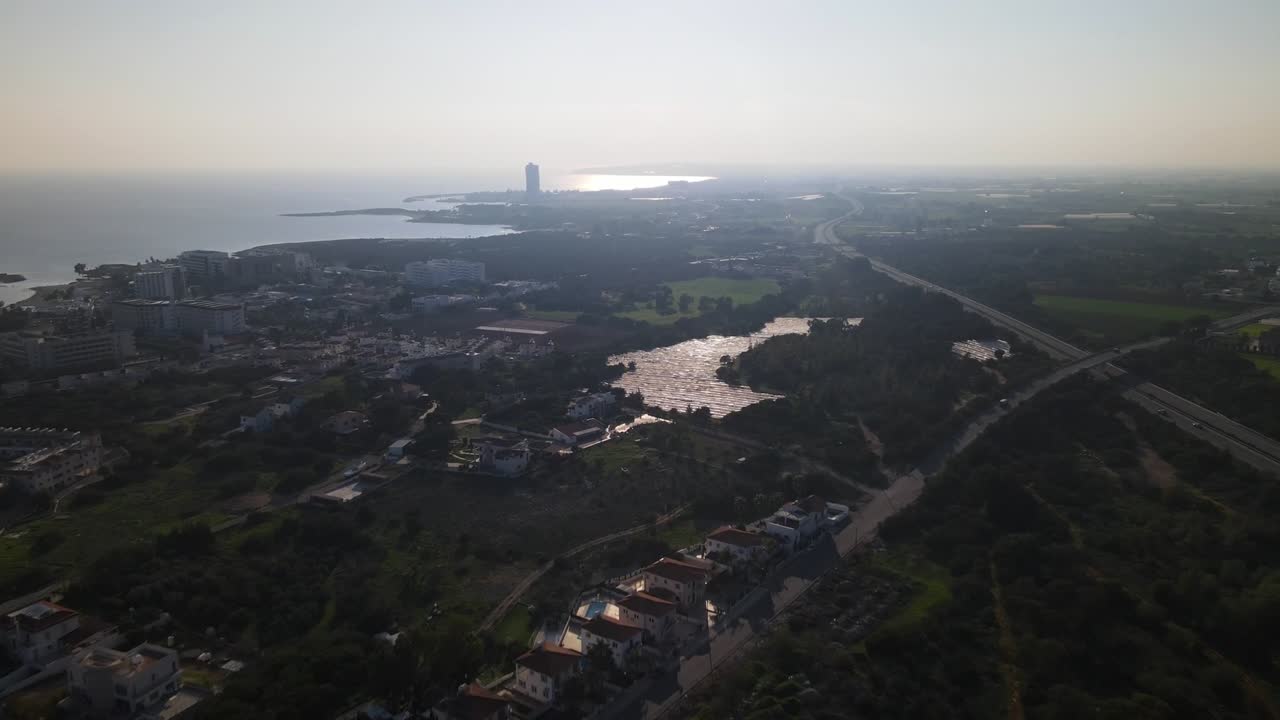 Drone aerial of Naples, Italy, showing a coastal city with a modern waterfront tower, highway cutting through green landscapes, and shimmering reflections on water leading to the horizon