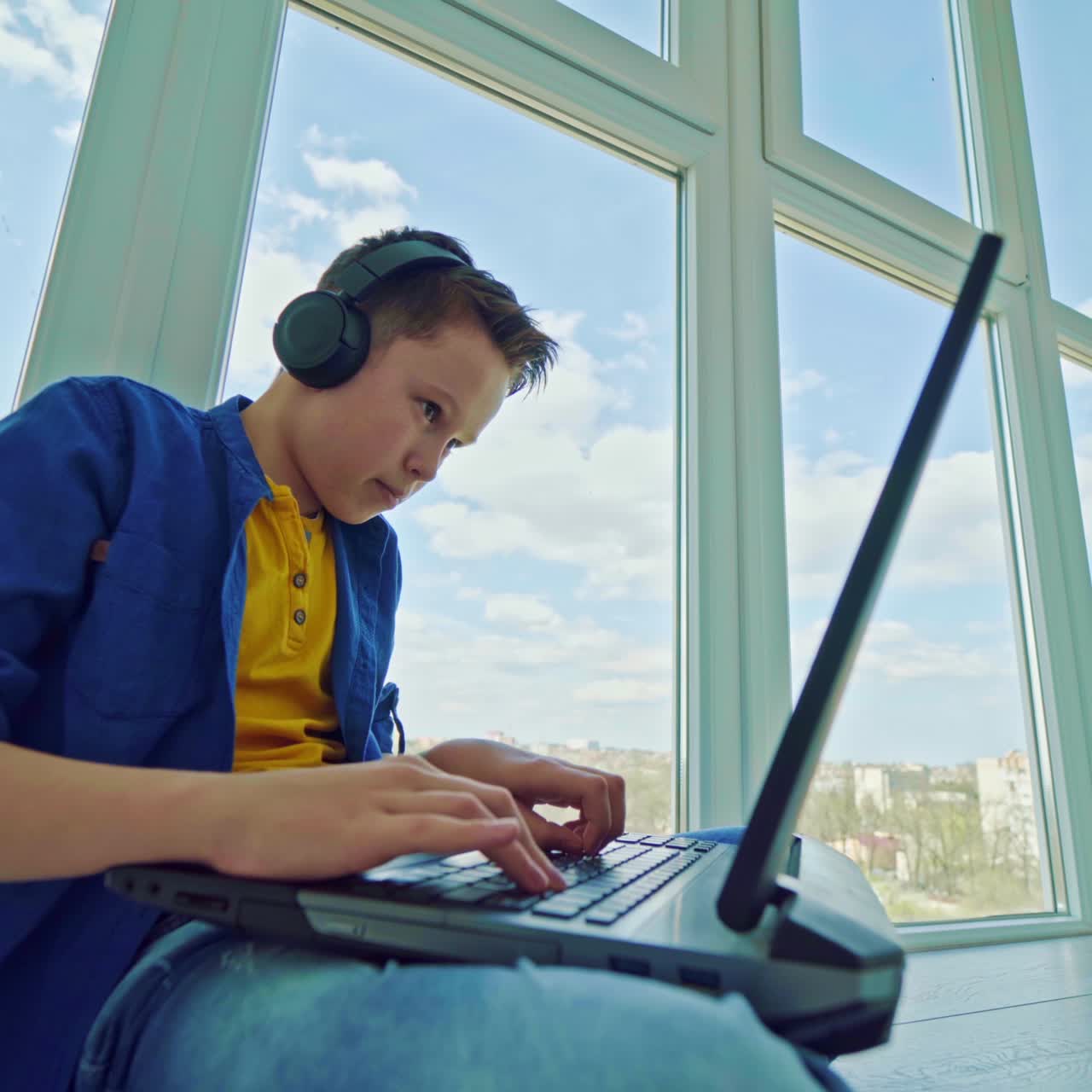 Teenager in headphones with a laptop by the window. Concentrated boy pressing buttons on a laptop while playing at home. Leisure time.