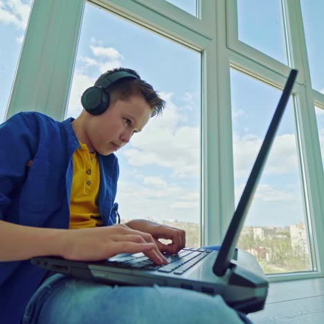 teenager in headphones with a laptop by the window. concentrated boy pressing buttons on a laptop while playing at home. leisure time.