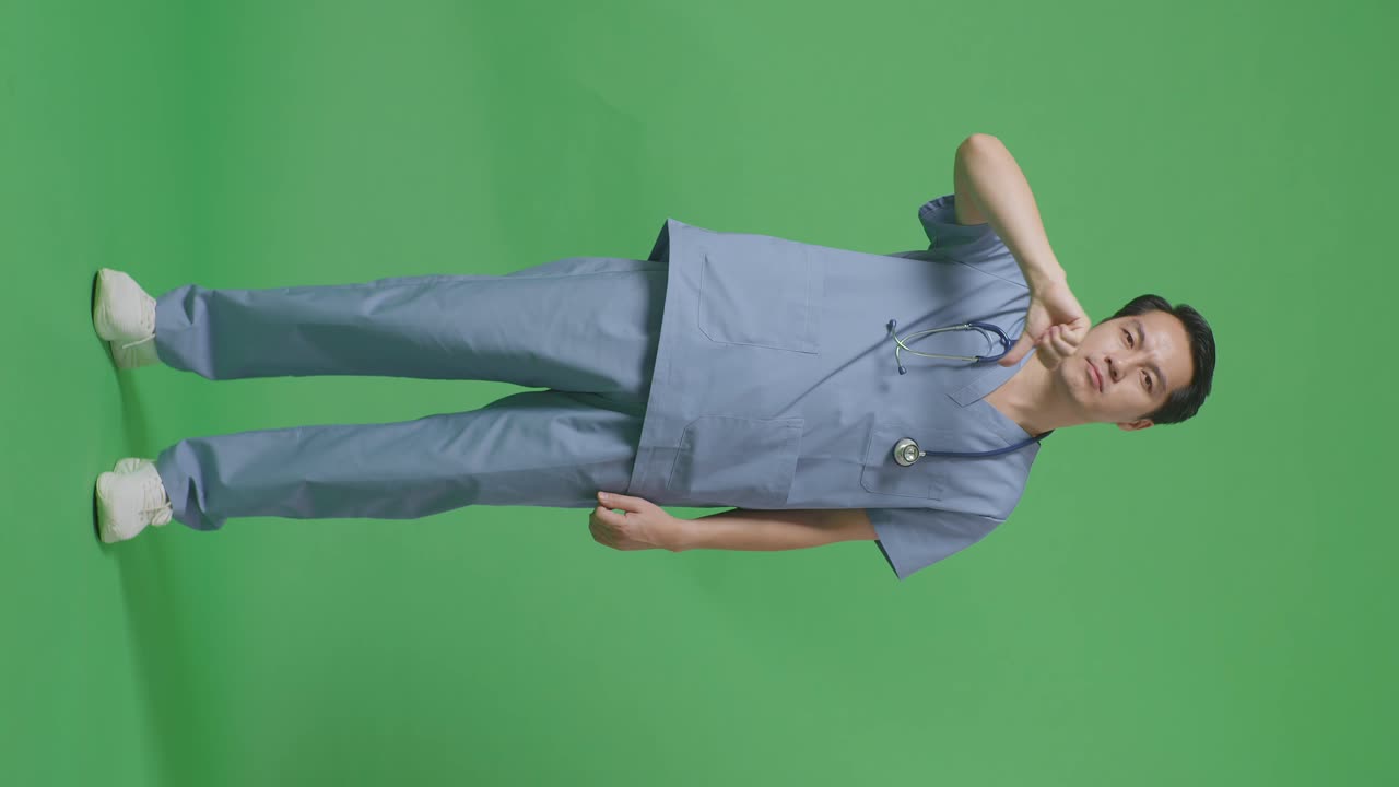 스테토스코프를 들고 있는 아시아 남성 의사의 몸 전체가 병원의 녹색 스크린 배경에 서 있는 동안 엄지손가락을 아래로 는 몸짓을 보여줍니다.
