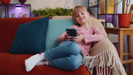 Child-toddler-hold-smartphone-watching-funny-cartoons,-chatting-with-friends-on-couch-alone-at-home