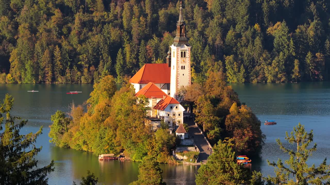 Widok z lotu ptaka na jezioro Bled z Cerkev Marijinega Vnebovzetja i kolorowymi drzewami w lesie. Wioślarze na łodziach