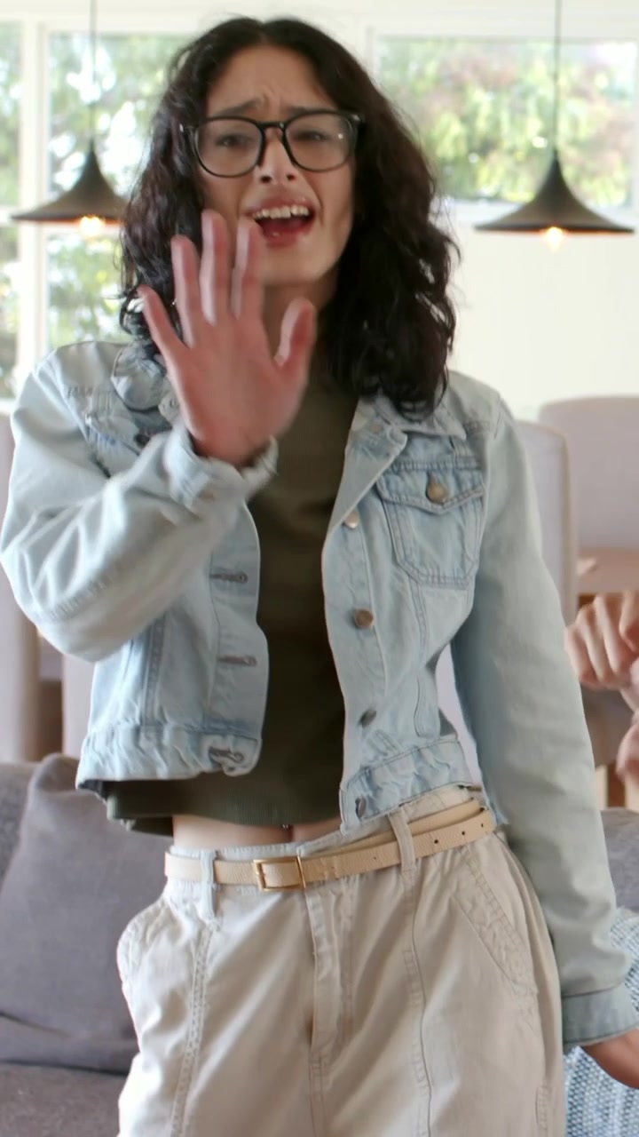 Donna che ondeggia con giacca di jeans e occhiali, facendo lip sync in soggiorno vicino al divano e alle finestre