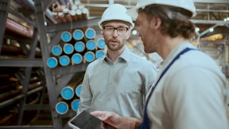 engineer and technician talking near stacked pipes in industrial warehouse