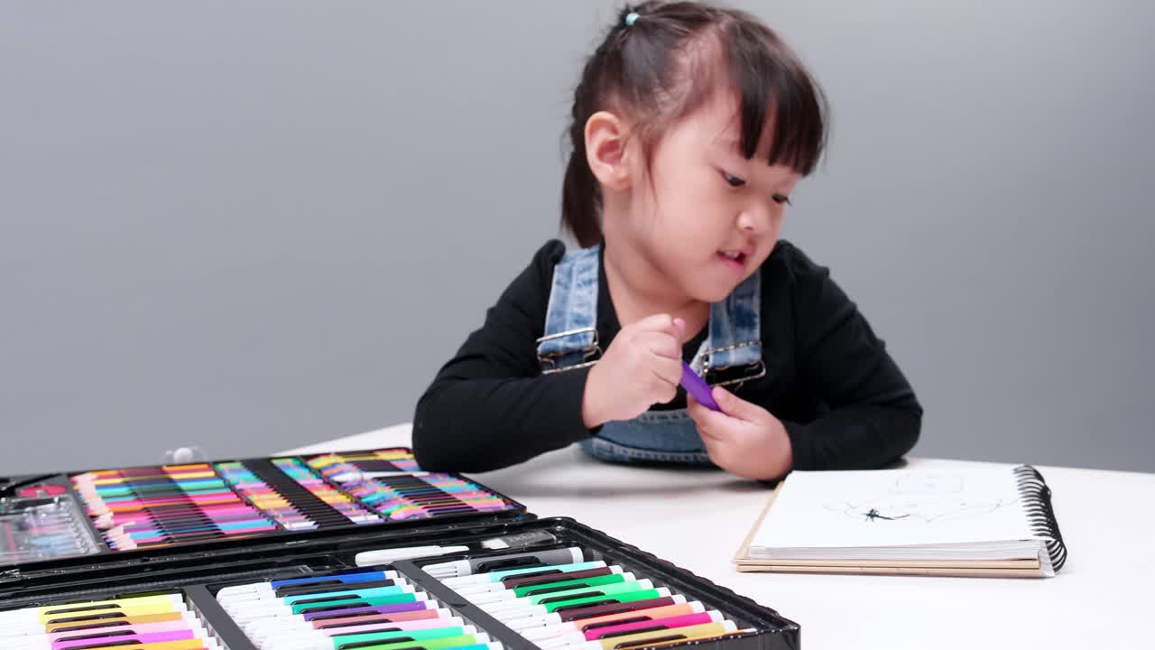 Una bambina asiatica si concentra sulla scelta di una penna magica colorata per dipingere un disegno su un quaderno.