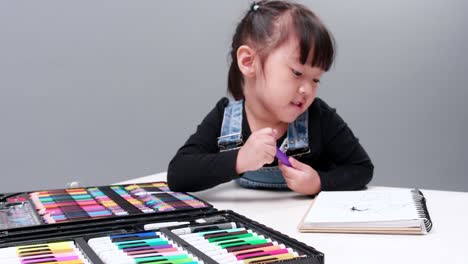 Una-Pequeña-Niña-Asiática-Se-Concentra-En-Elegir-Un-Bolígrafo-Mágico-De-Color-Para-Pintar-Una-Imagen-En-Un-Cuaderno