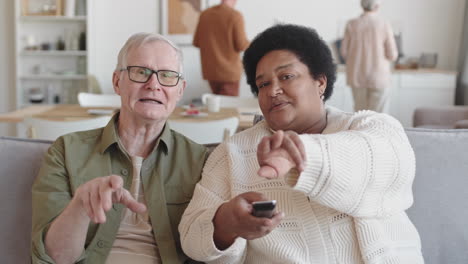 waist up pov of grey-haired caucasian man and african woman sitting on sofa at home, watching invisible tv, discussing it, smiling, pointing fingers