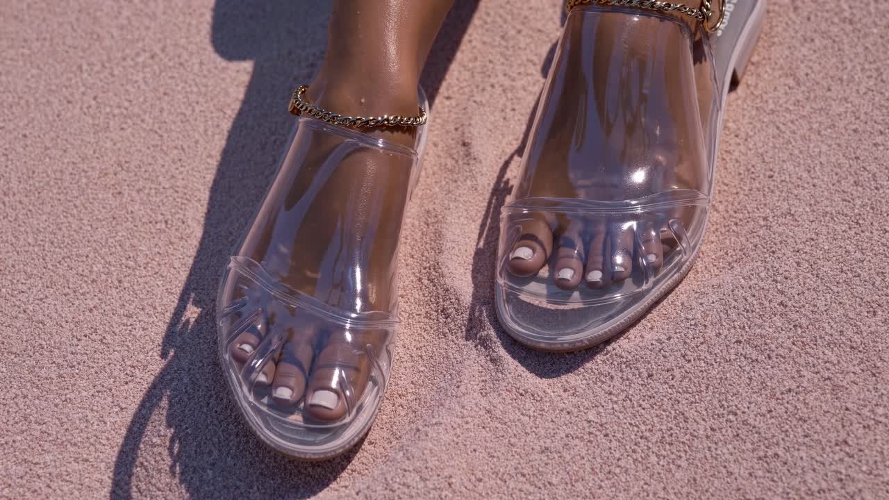 Close up of woman's feet wearing stylish transparent jelly sandals with gold chain anklet on a beautiful pink sand beach, enjoying a relaxing summer day