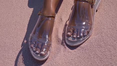 close up of woman's feet wearing stylish transparent jelly sandals with gold chain anklet on a beautiful pink sand beach, enjoying a relaxing summer day