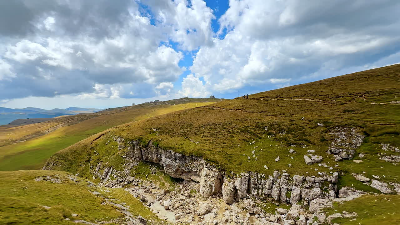 Verdi colline e paesaggio roccioso vicino a catene montuose con nuvole nel cielo. Persone camminano su un verde pendio vicino a un'area rocciosa sotto un cielo blu con nuvole.