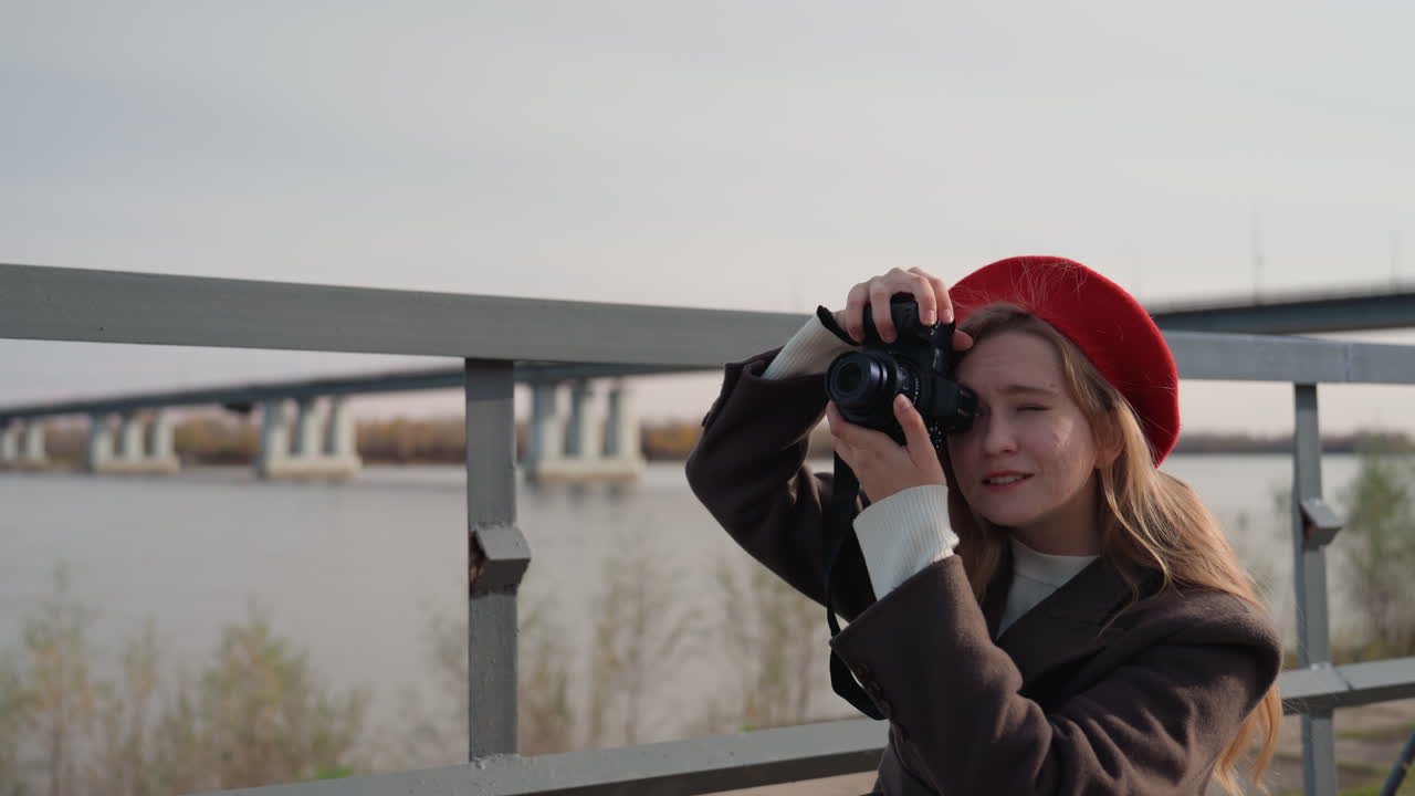 Fotografa caucasica in riva al fiume, seduta su una panchina con macchina fotografica vintage e berretto rosso. Sguardo contemplativo verso il ponte, luce autunnale smorzata, atmosfera calma sul lungofiume, tavolozza di colori tenui.