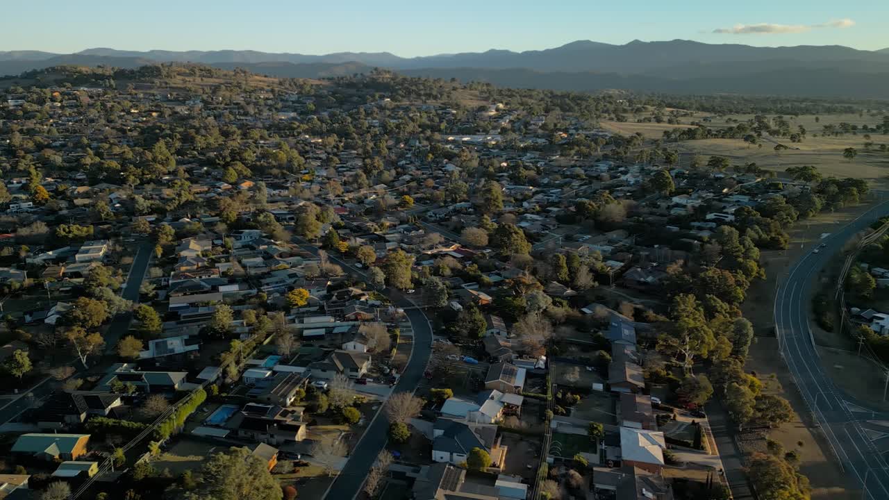 Movimiento aéreo lateral a través de las frondosas calles y tejados de Rivett, mientras el sol proyecta largas sombras doradas sobre el suburbio