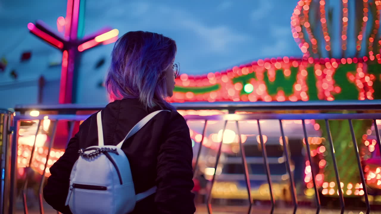 cinemagraph de una adolescente caucásica con mochila mirando por encima de la valla en la feria de atracciones a la luz de la noche