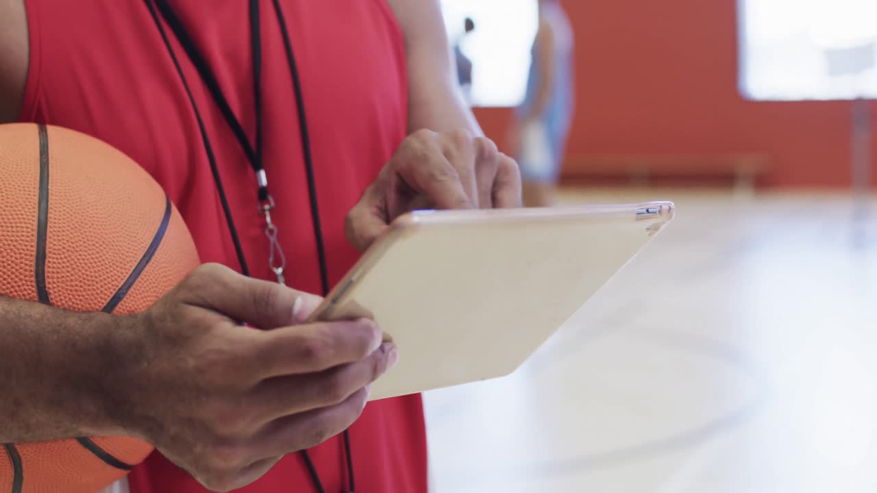 sección media de un entrenador de baloncesto masculino biracial con una pelota usando una tableta en una cancha cubierta, en cámara lenta