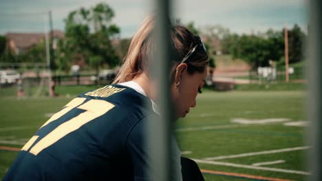 female athlete is looking intensely down as she laces her shoes while sitting on a row of bleachers in front of a sports field