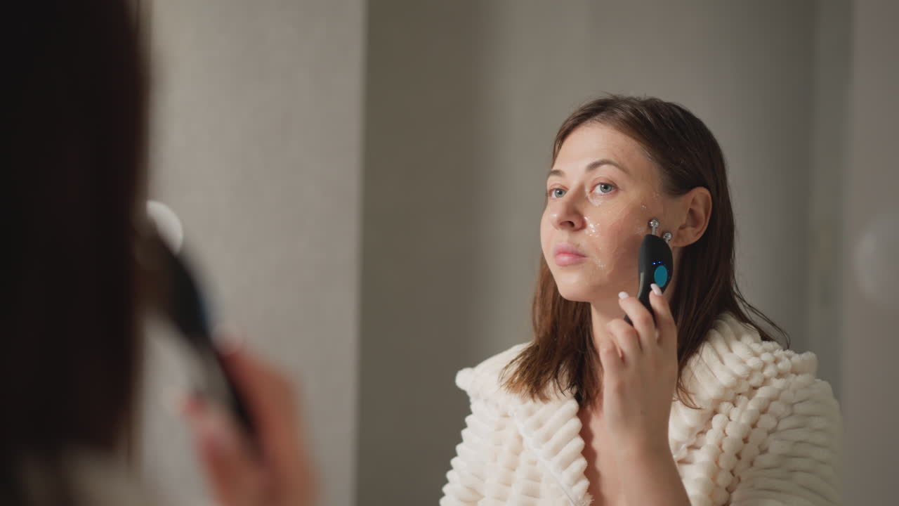 Female does microcurrent procedure for face lift in bathroom. Attractive brunette woman in robe does facial treatment using microcurrent slow motion