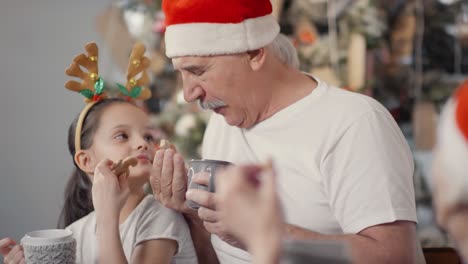 Happy-Grandfather-And-Cute-Little-Girl-In-Santa-Hat-And-Festive-Headband-Drinking-Hot-Chocolate-And-Eating-Cookies-While-Chatting-With-Grandmother-On-Christmas-Eve