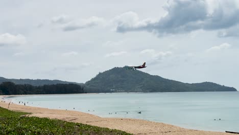 an airplane approaches for landing over mai khao beach, phuket, with serene ocean and mountain views under cloudy skies