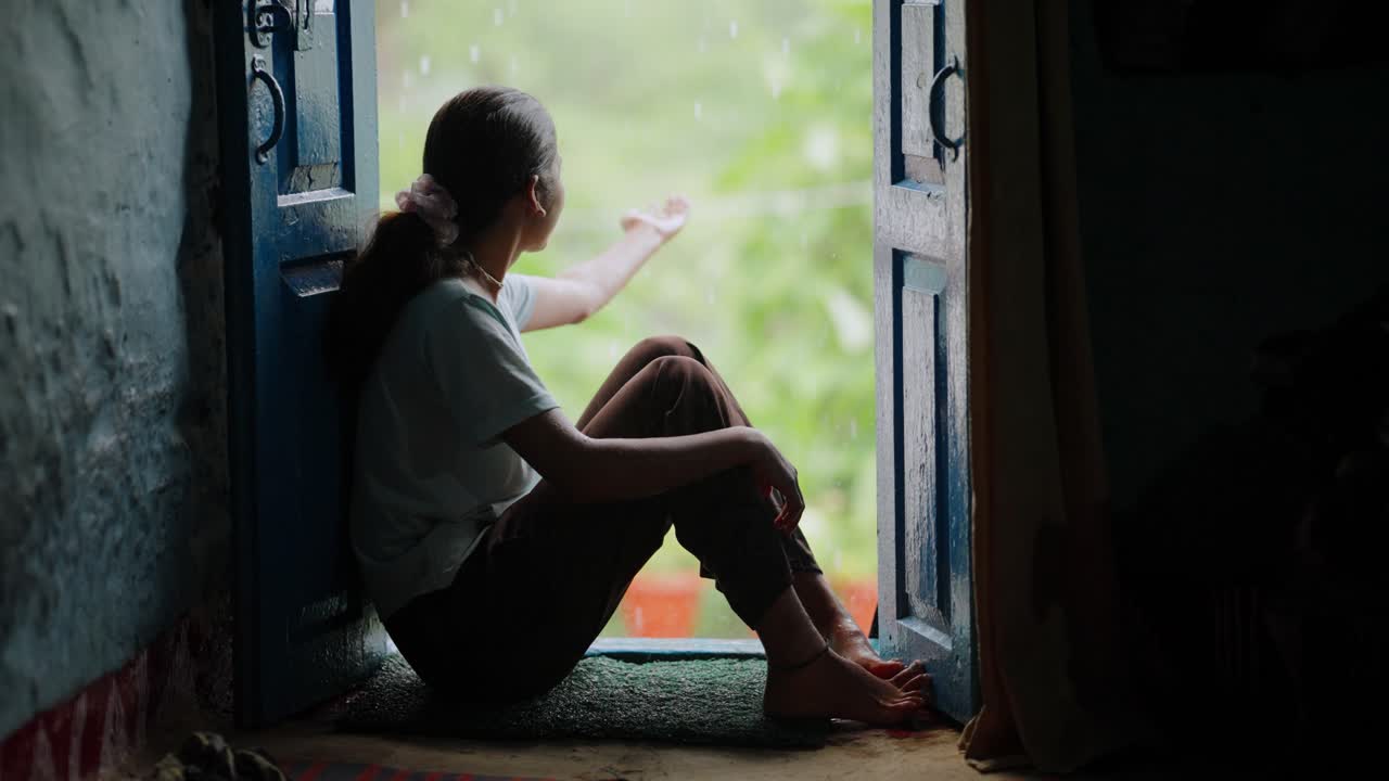Indian girl in traditional clothing enjoying rain from her home entrance, peaceful village life, 4k video
