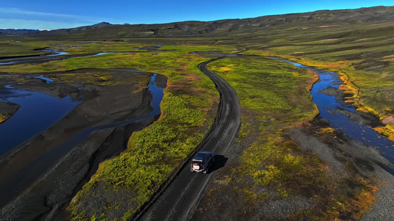 Ripresa aerea con drone di un SUV solitario che percorre una tortuosa strada sterrata attraverso gli altopiani islandesi, con fiumi intrecciati blu, pianure muschiose e un vasto terreno vulcanico sotto cieli sereni.