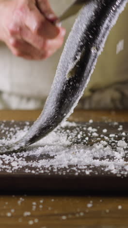 man cook uses knife to clean large fat pickled herring from salt on wooden board at kitchen table close view. healthy nutrition of seafood