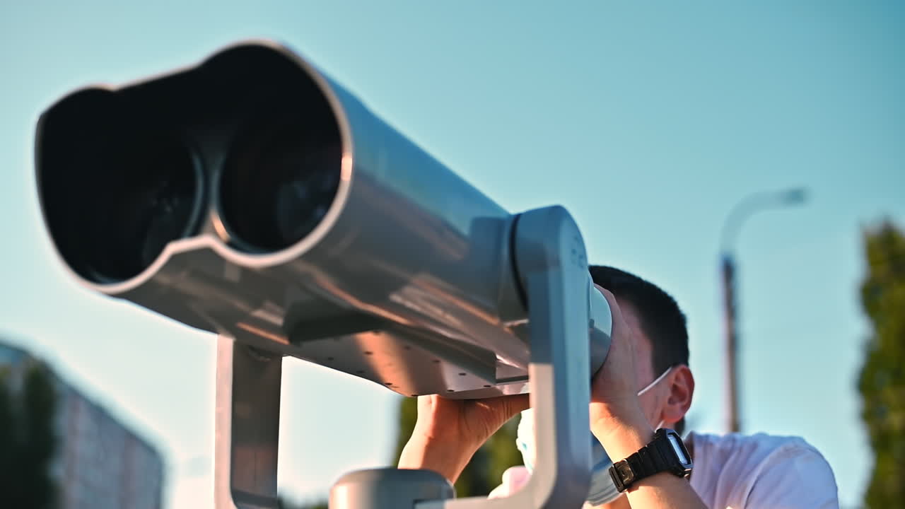 Young man in protective medical mask and white t-shirt looking into a tower viewer with city and green trees on background. Sunny day. Corona Virus idea. Safe travel during the pandemic. Slow motion