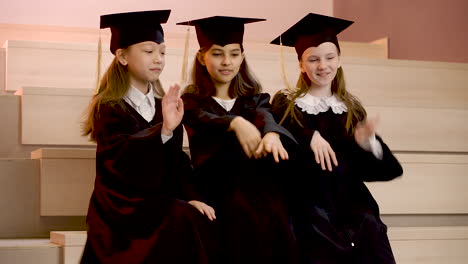 tres niñas felices con toga y birrete jugando juntas mientras se sientan en las escaleras en la ceremonia de graduación de preescolar