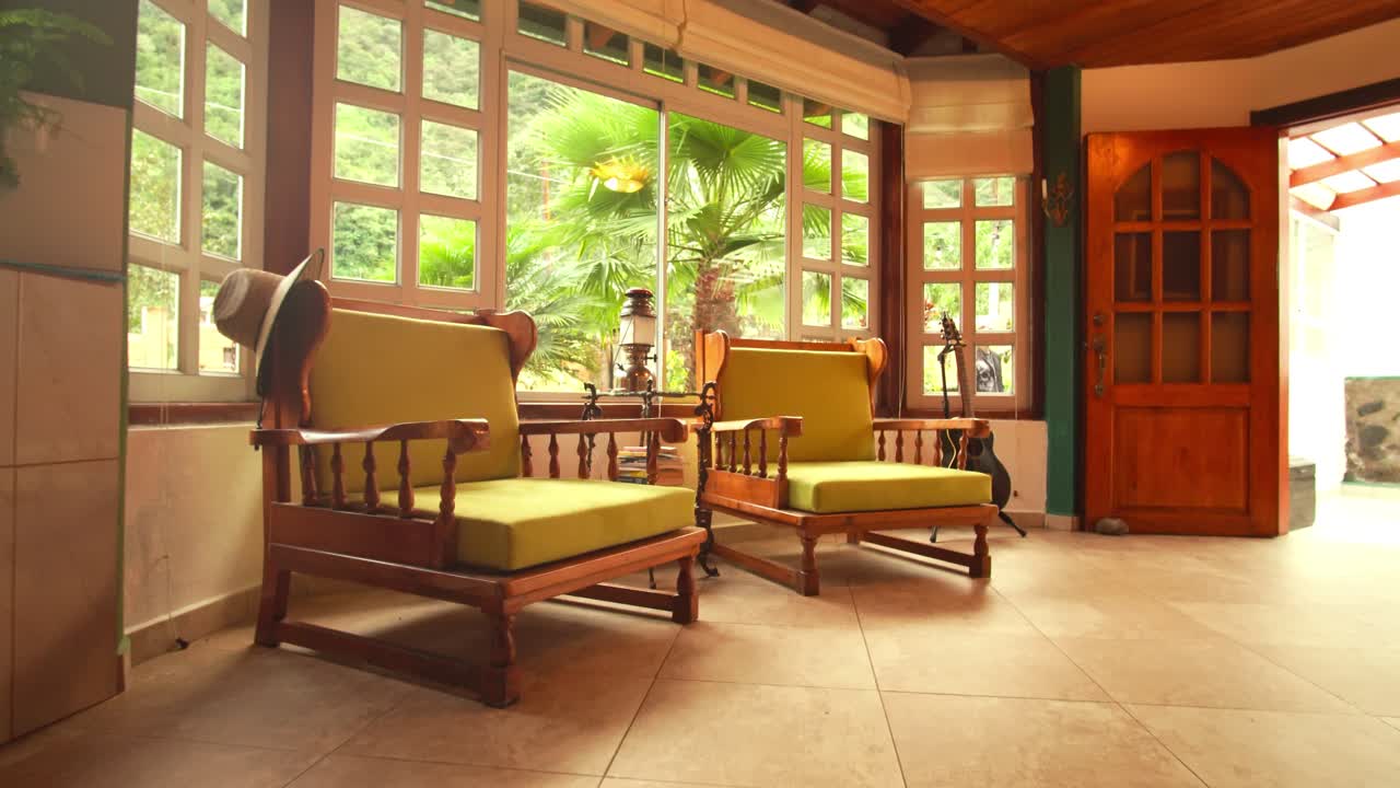 Dolly in on a rustic-modern living room with wooden chairs, tiled floor and large windows looking out to tropical greenery.