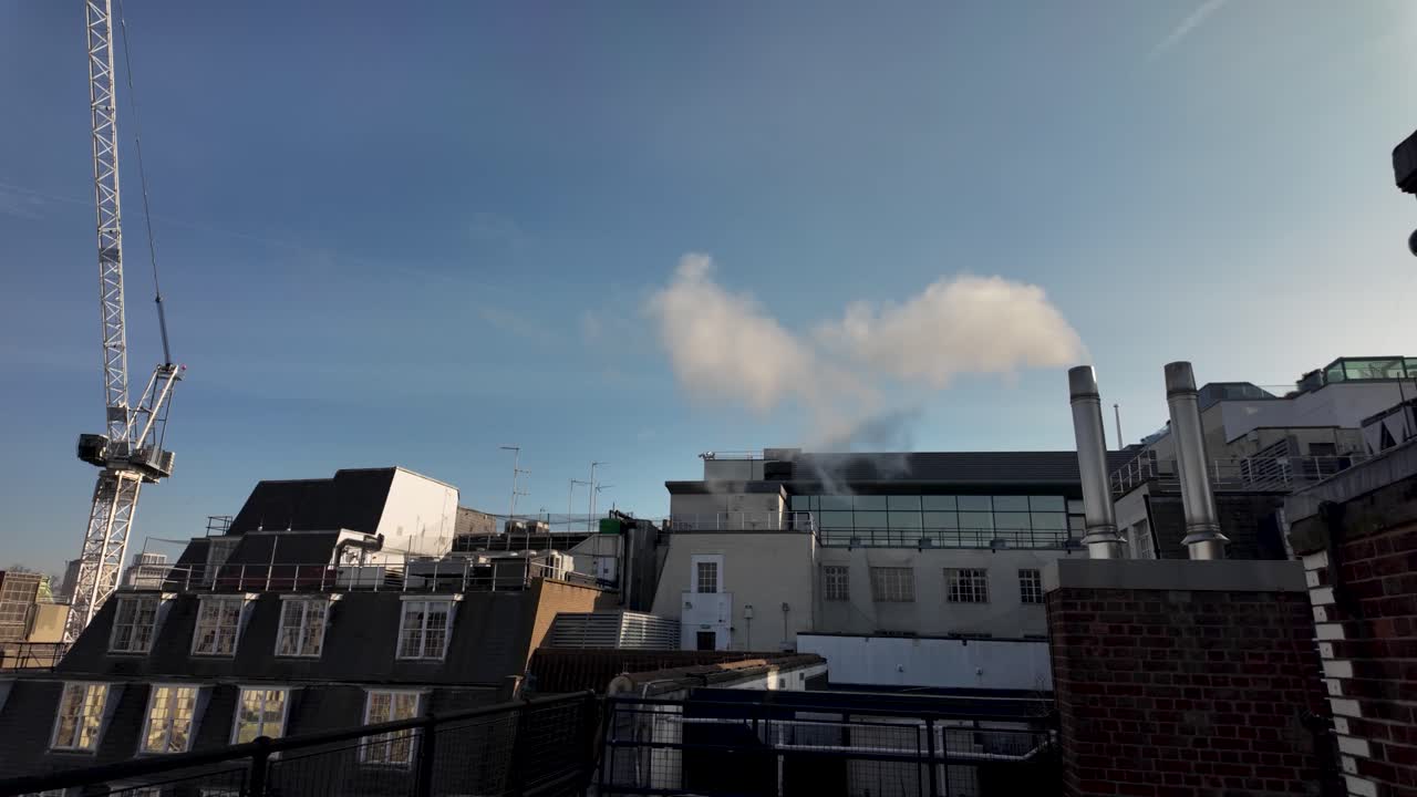 White smoke is coming out of metal chimney flues on a rooftop of a London building, with a crane and blue sky in the background