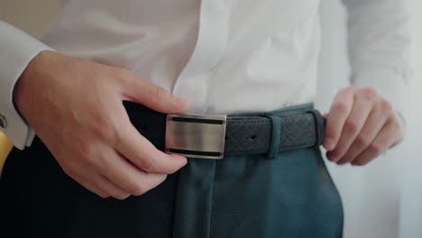 man adjusting a black belt on his trousers, dressed in a white shirt, preparing for a formal event