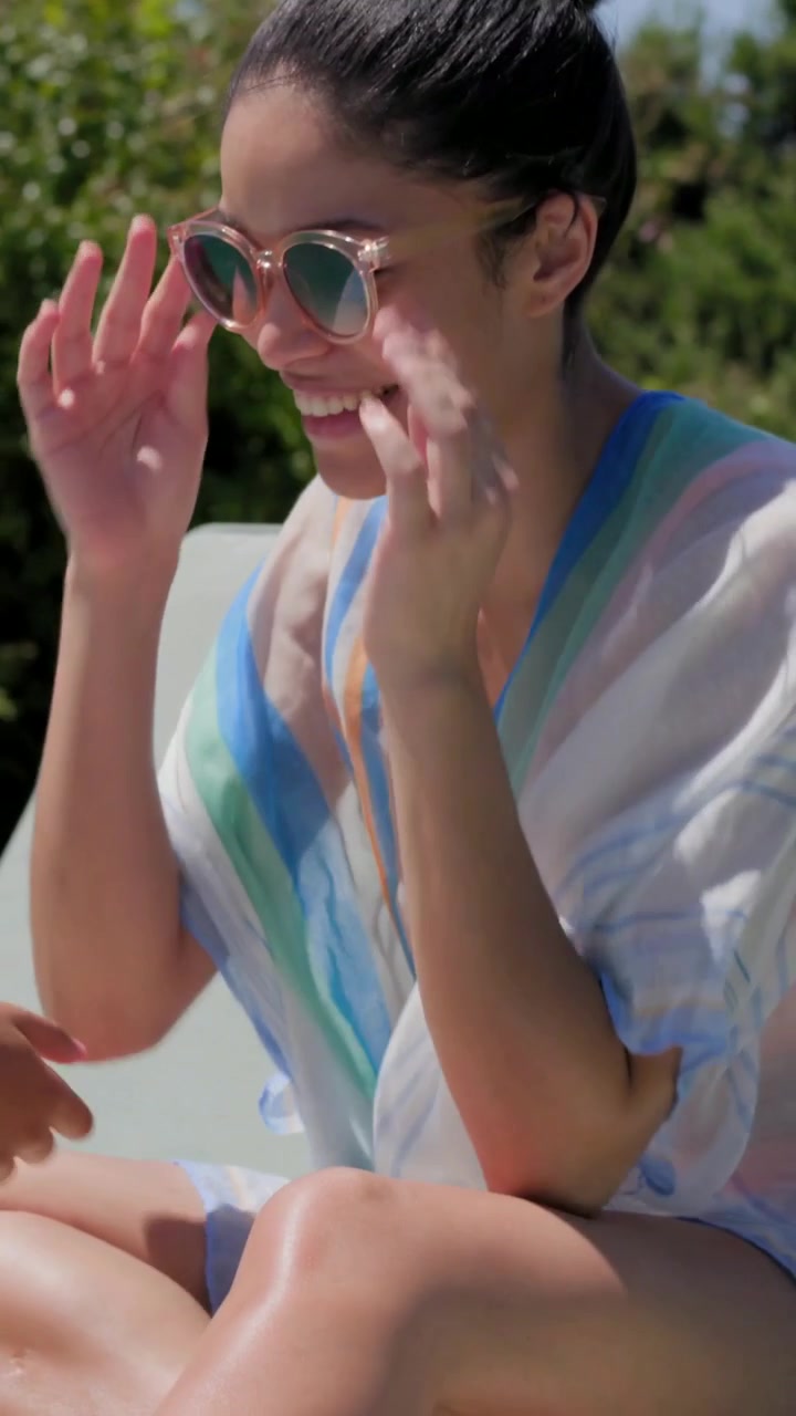 Vertical video: Reaching woman to offscreen hand, rubbing sunscreen on deck for sun care in kaftan