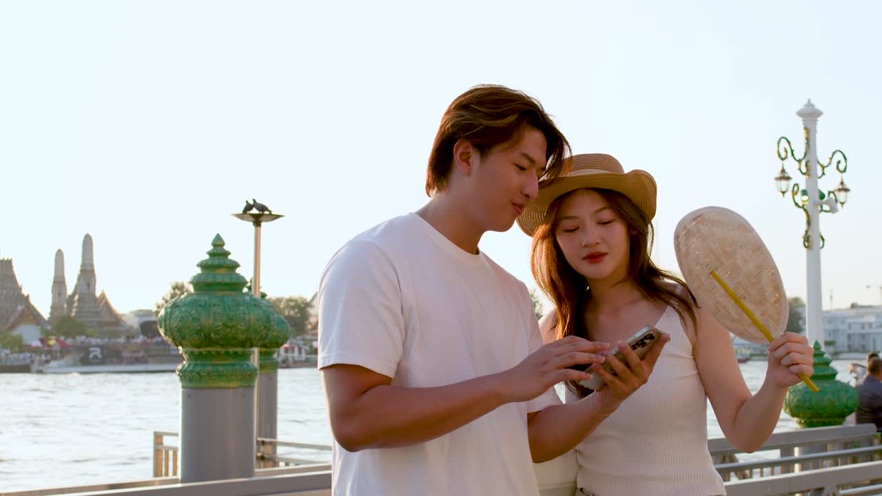 Young couple checks smartphone together near riverside temple, warm sunset lighting, relaxed vacation mood