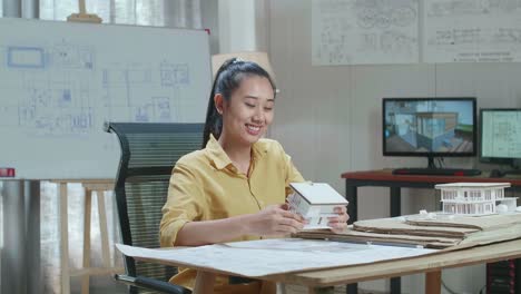 asian woman engineer holding paper model of house warmly smiling and showing thumb up to camera at the office