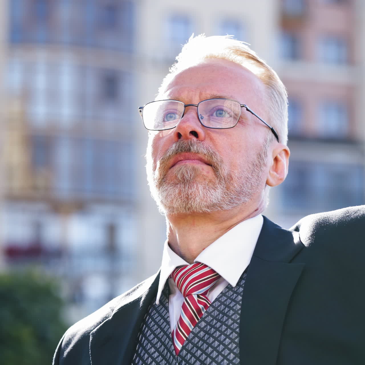 Close-up portrait of a senior man with grey hair with beard sitting on the city background. Old businessman in eyeglasses straighten his tie and looks to camera.