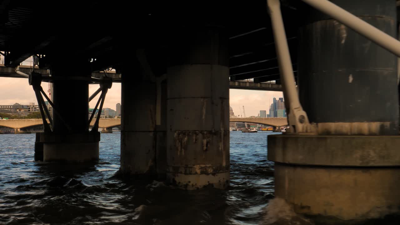 Floating on the river Thames in London on a boat, passing underneath a modern bridge full of people and tourists, passing by the skyscrapers of the business district in central London on a sunny day.