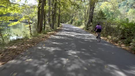 camera moving along with and catching up to a woman biking on a shady rural road with a river to her left side