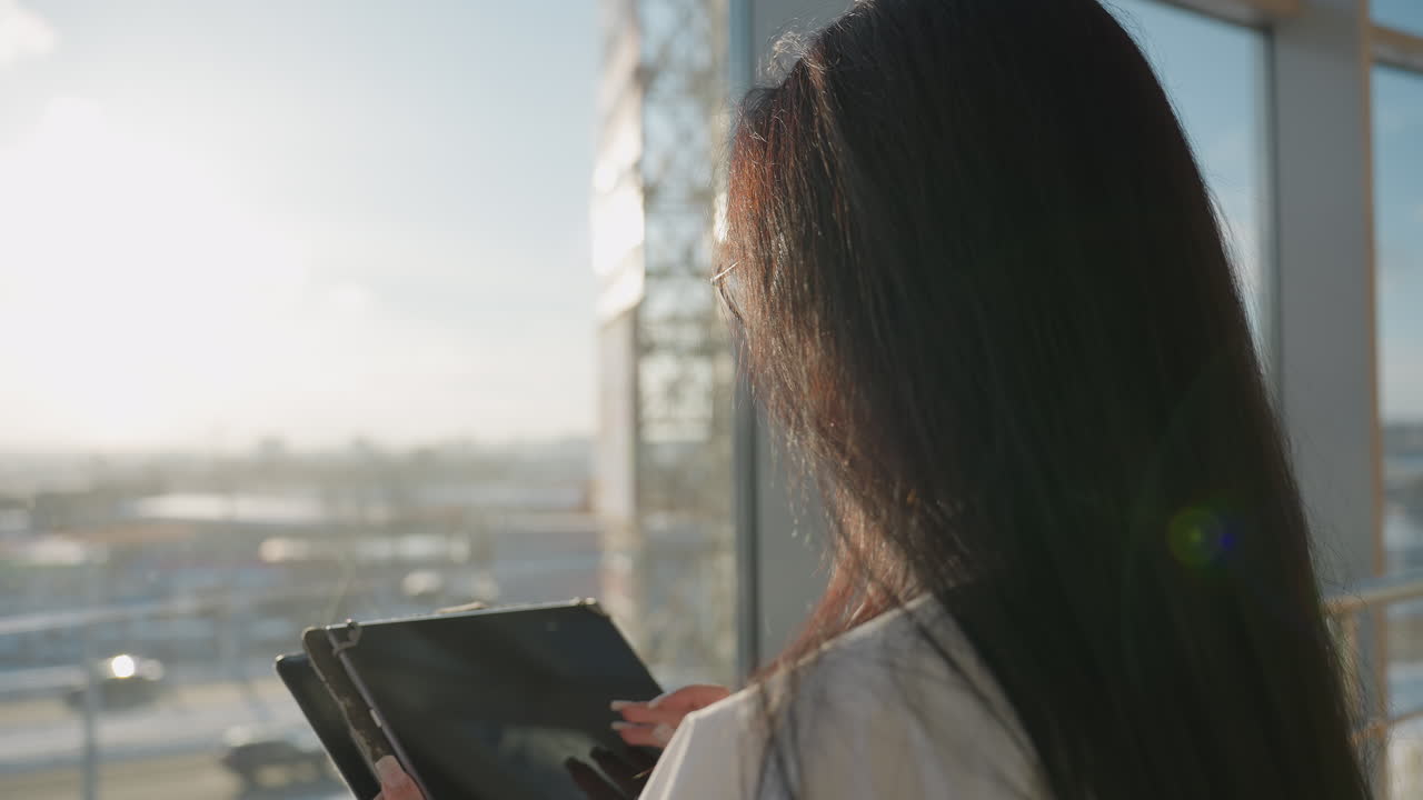 Close up of professional woman adjusting glasses while holding digital tablet near glass window, sunlight shining through, with blurry outdoor view of moving cars in urban environment