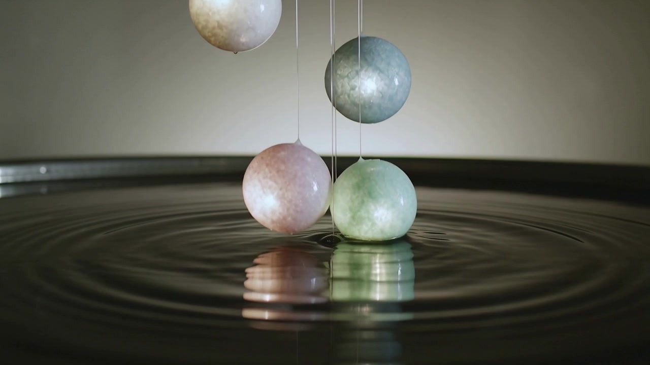 Lowering by thin strings, pastel spheres contacting round tray of dark liquid, producing ripples