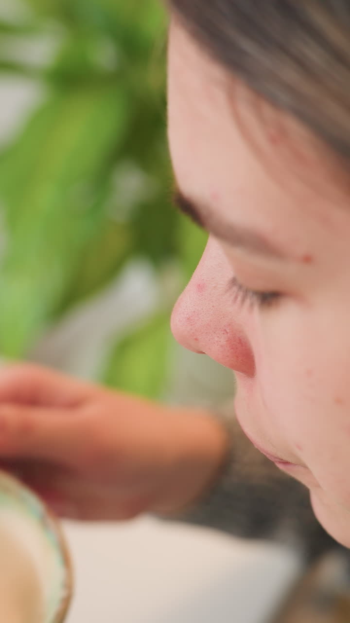 Mujer caucásica sosteniendo una taza color menta con espuma de latte, smartphone en el escritorio y planta de fondo; manos sosteniendo un plato de cerámica caliente, primeros planos de sorbo y perfil mostrando pecas y acné, breve pausa para teletrabajar