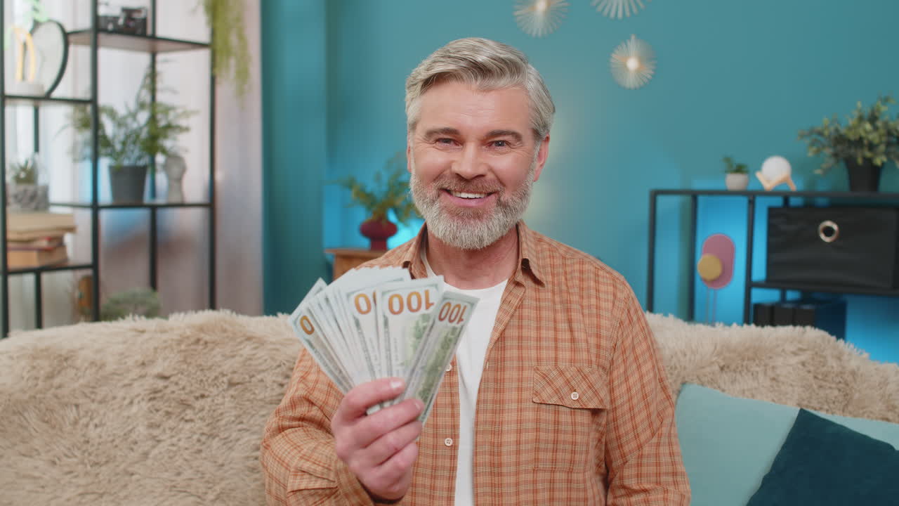 Mature man sitting home with smartphone and dollars money cash celebrating successful money transfer
