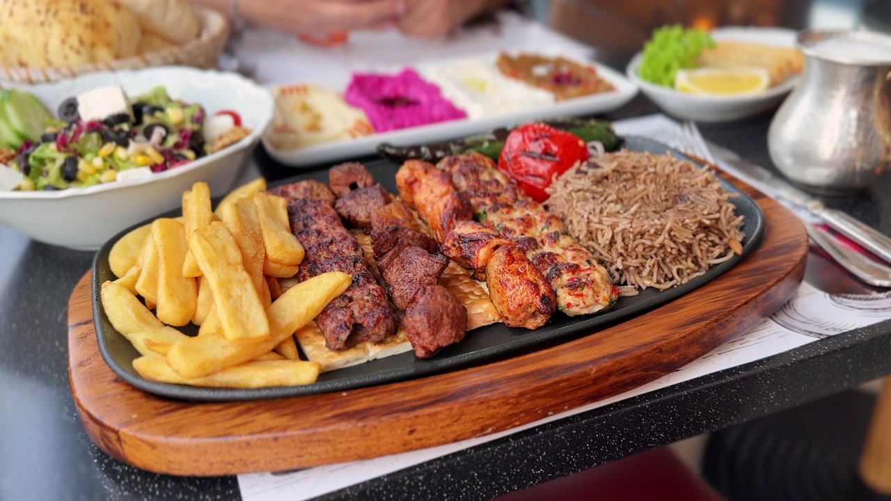 Abbondante piatto di grigliata mista mediorientale servito con patatine fritte, riso, insalata fresca e salse tradizionali su un tagliere di legno su un tavolo luminoso di un ristorante durante il giorno