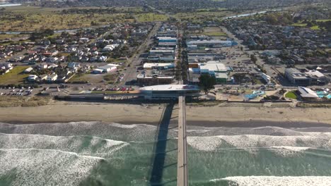 Excelente-Vista-Aérea-Del-Paisaje-Urbano-Costero-Del-Océano-Ondeando-Una-Larga-Playa-De-Arena,-Zona-De-Viviendas-Junto-Al-Mar-Y-Gente-Caminando-En-El-Muelle