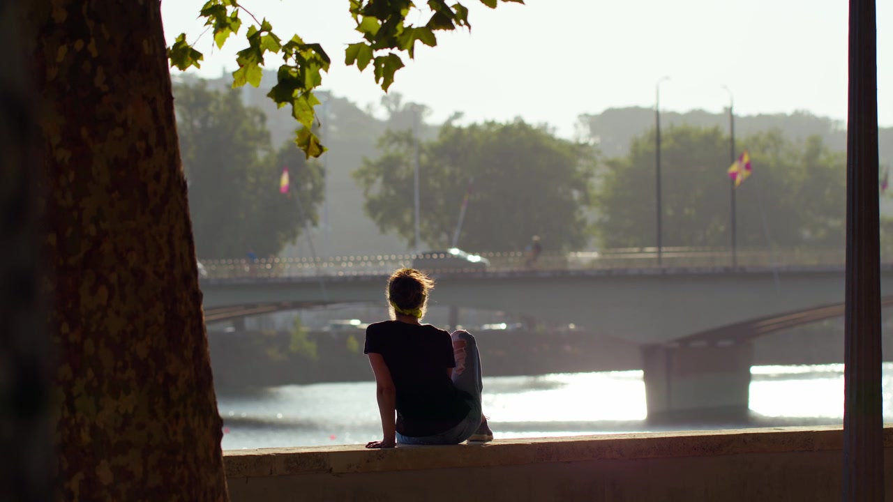 Silhouette of a girl watching the sunset by the Mondego River, with wind blowing her hair and tree leaves, Santa Clara Bridge in the background, Coimbra