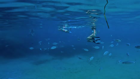 Shoal-of-fish-swims-close-to-mother-and-daughter-snorkeling-underwater-in-deep-blue-sea