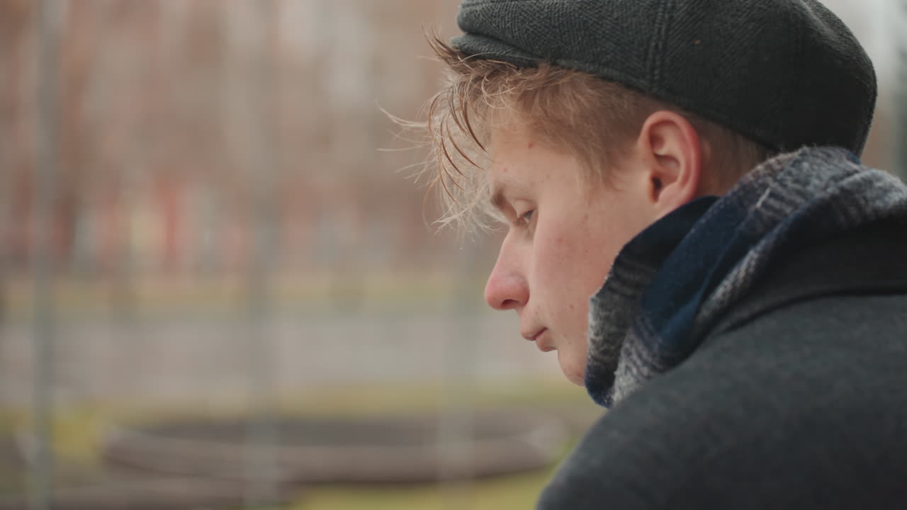 Bílý tvůrce otáčí hlavu k fotoaparátu u zábradlí na nábřeží, studený dech a dešťové kapky, ostrý, soustředěný pohled zblízka, baret a šála, pozadí městského parku, intenzivní výraz, nezávislý režisér a model.