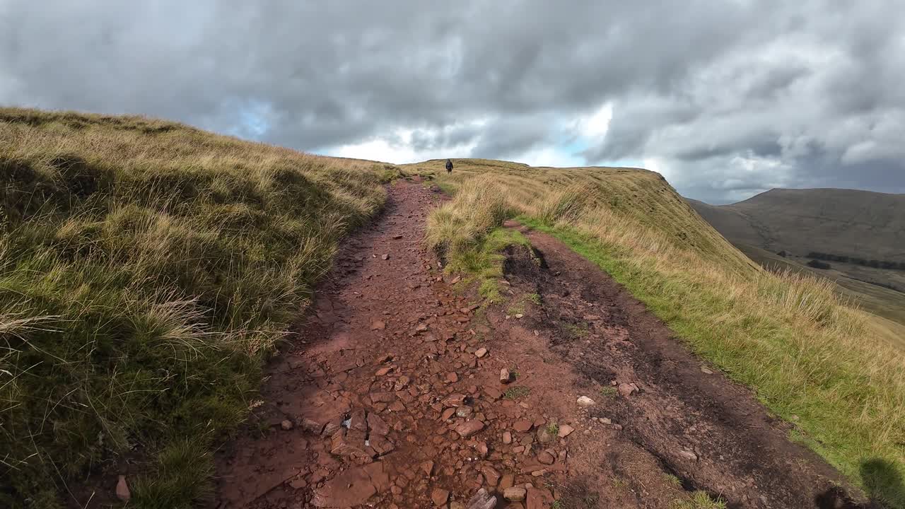 A rugged mountain trail winds through the Brecon Beacons National Park, leading toward the Pen y Fan summit under dramatic skies, showcasing wild grass, rocky paths, and rolling Welsh hills
