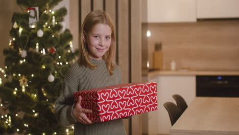 vista frontal de una chica rubia con suéter verde sosteniendo regalos en una habitación decorada con un árbol de navidad