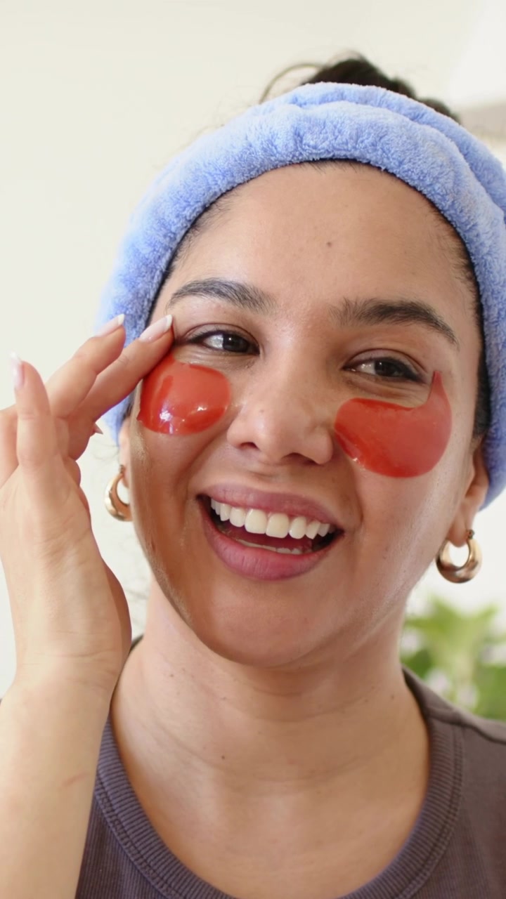 Vertical video: Noticing patch slip, woman smoothing orange eye patches at home, with blue headband