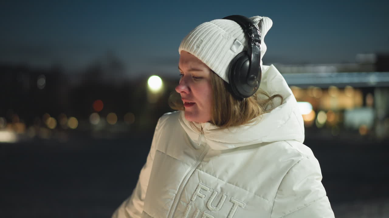 Cheerful young lady moving body in rhythm to music under bright park lights on snow covered walkway at night wearing white coat beanie and headphones surrounded by ambient urban winter scene